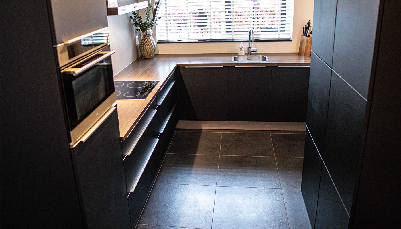 L-vormige keuken met zwarte gewrapte kasten en een warm houten werkblad bij het raam.
